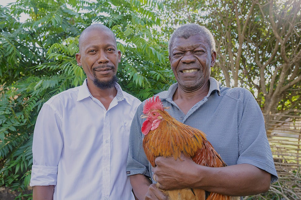 Two men stand outside in Zambia – one of them holds a chicken in his arms.