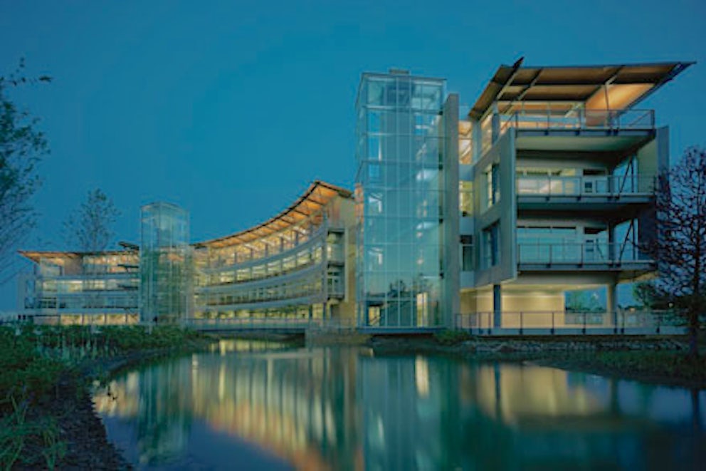 A view of the Heifer headquarters building at night, the lights reflecting off the surface of the retention pond that surrounds the building.
