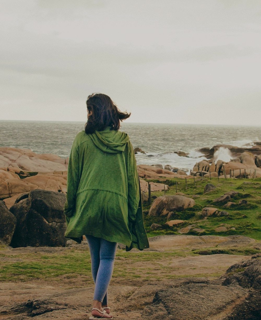A woman takes a walk on a rocky beach in a green jacket.