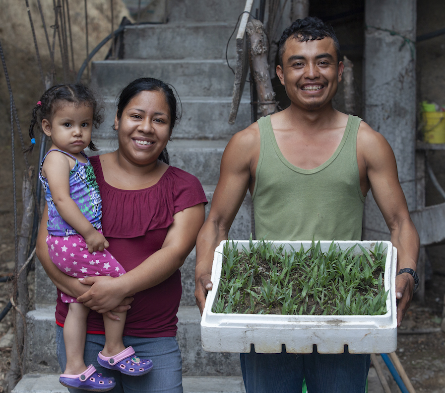 A couple stands, smiling, with the woman holding a young girl, and the man holding a container of agave seedlings.