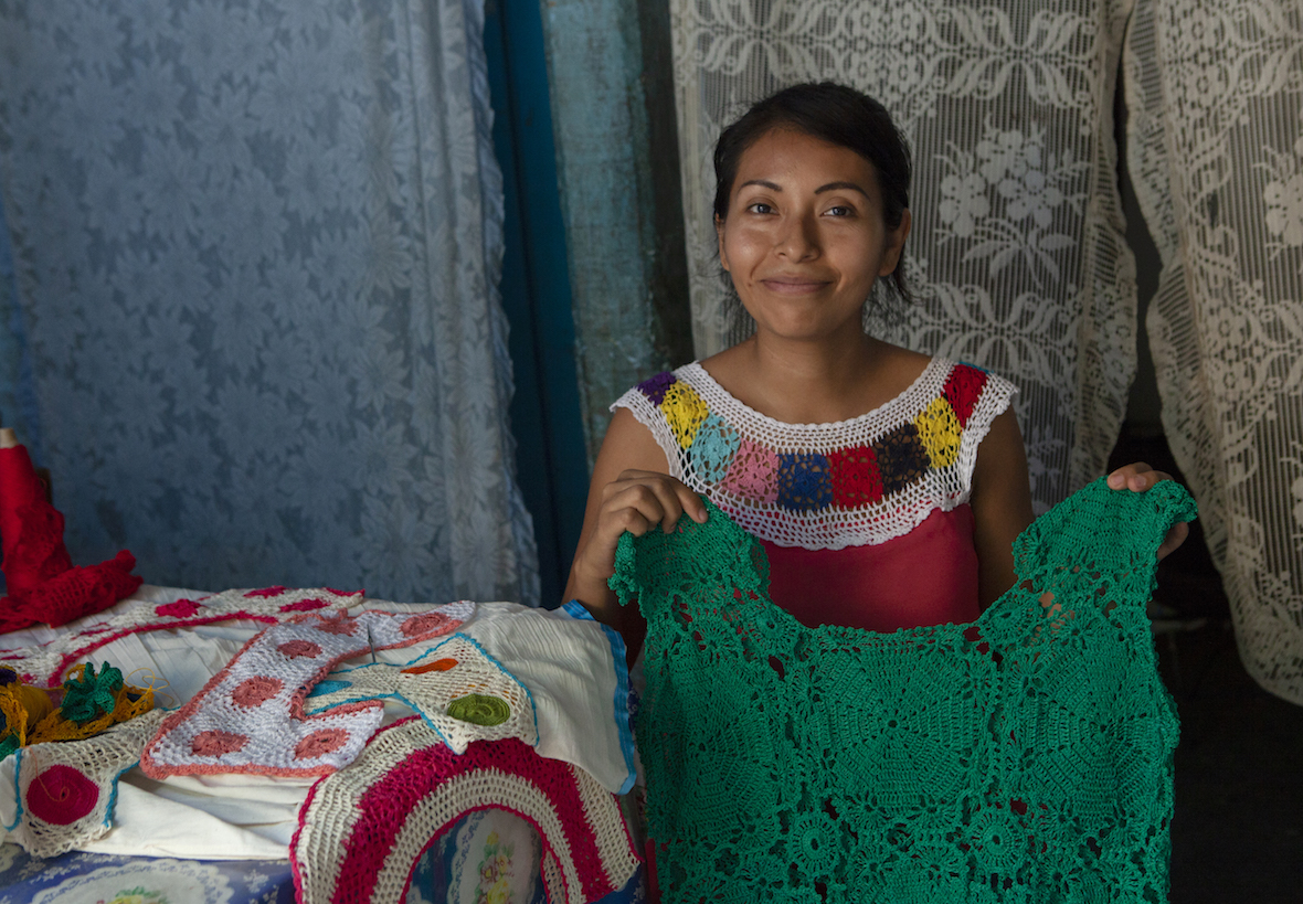 A young woman displays a green fabric she has made, with more hand made clothes on the bed beside her.
