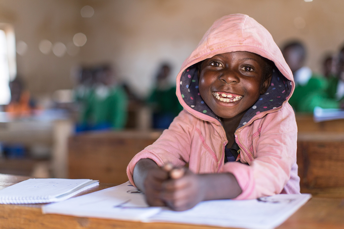 A smiling young boy sits in a classroom in a bright pink jacket.