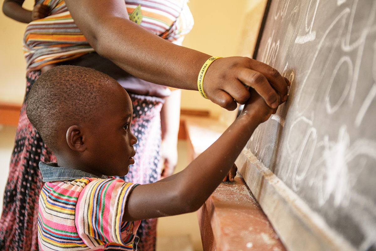 A teacher holds a student's hand and helps him write on a chalkboard.