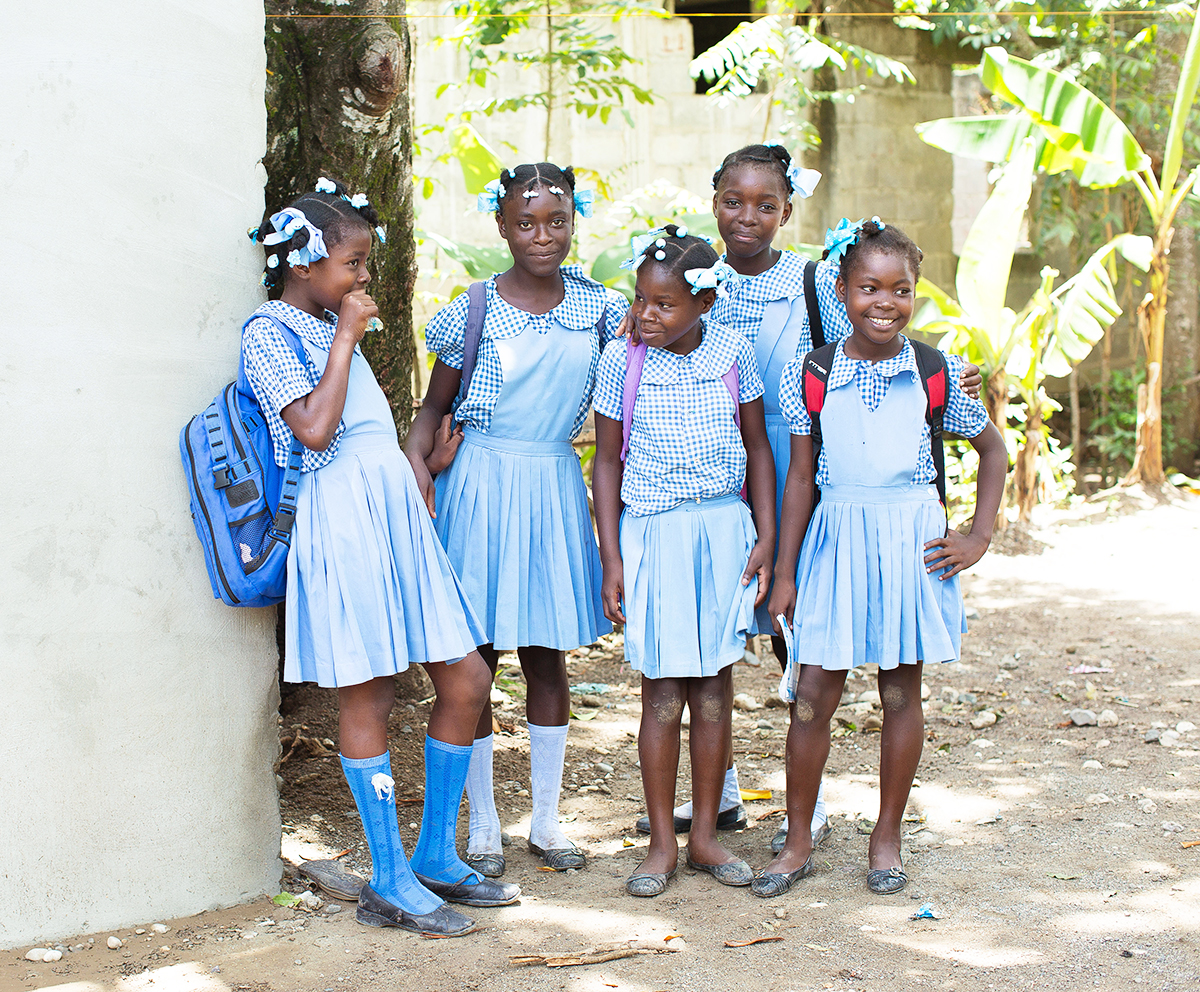 A group of 5 young students in their uniforms pose on a white wall outside.