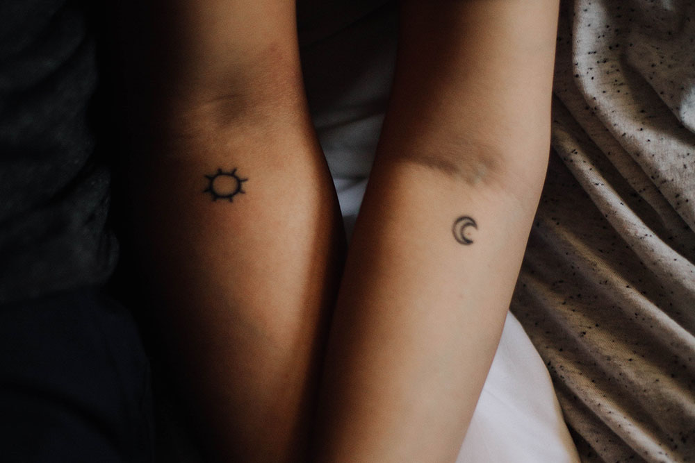 Two women's arms crossing, one with a sun tattoo and one with a moon tattoo.