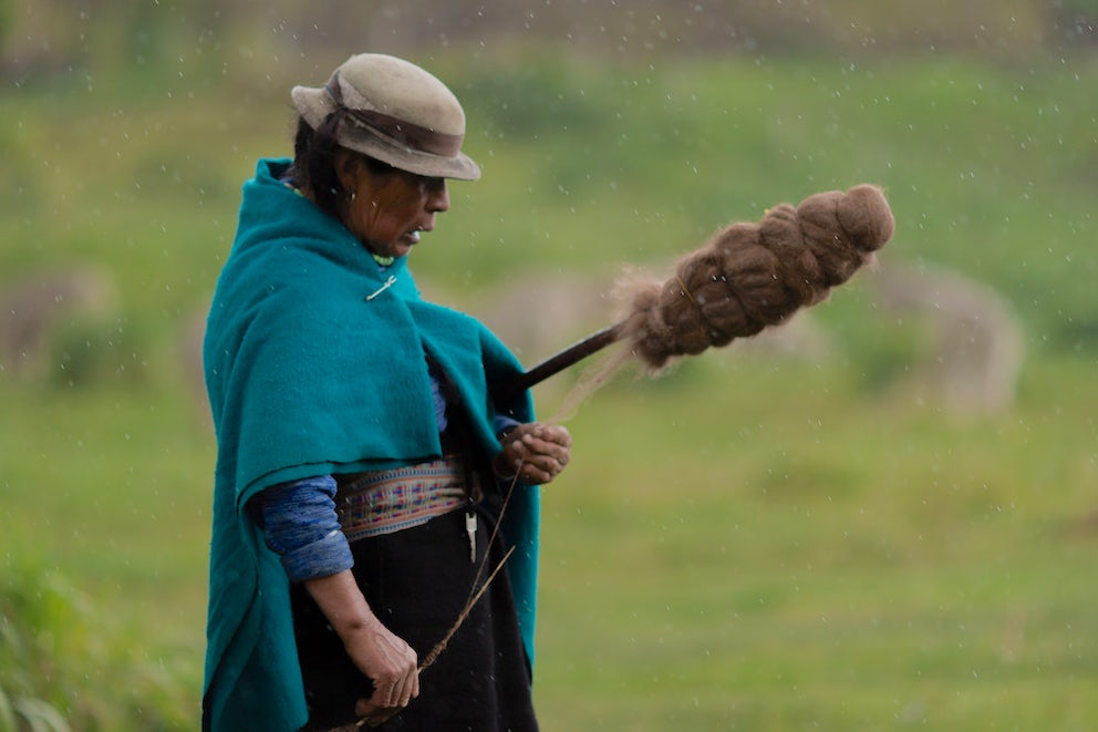 A member of the Tombohaushca, Ecuador community spins alpaca fiber in the drizzle.. Her colorful faja peeks out from underneath her poncho.