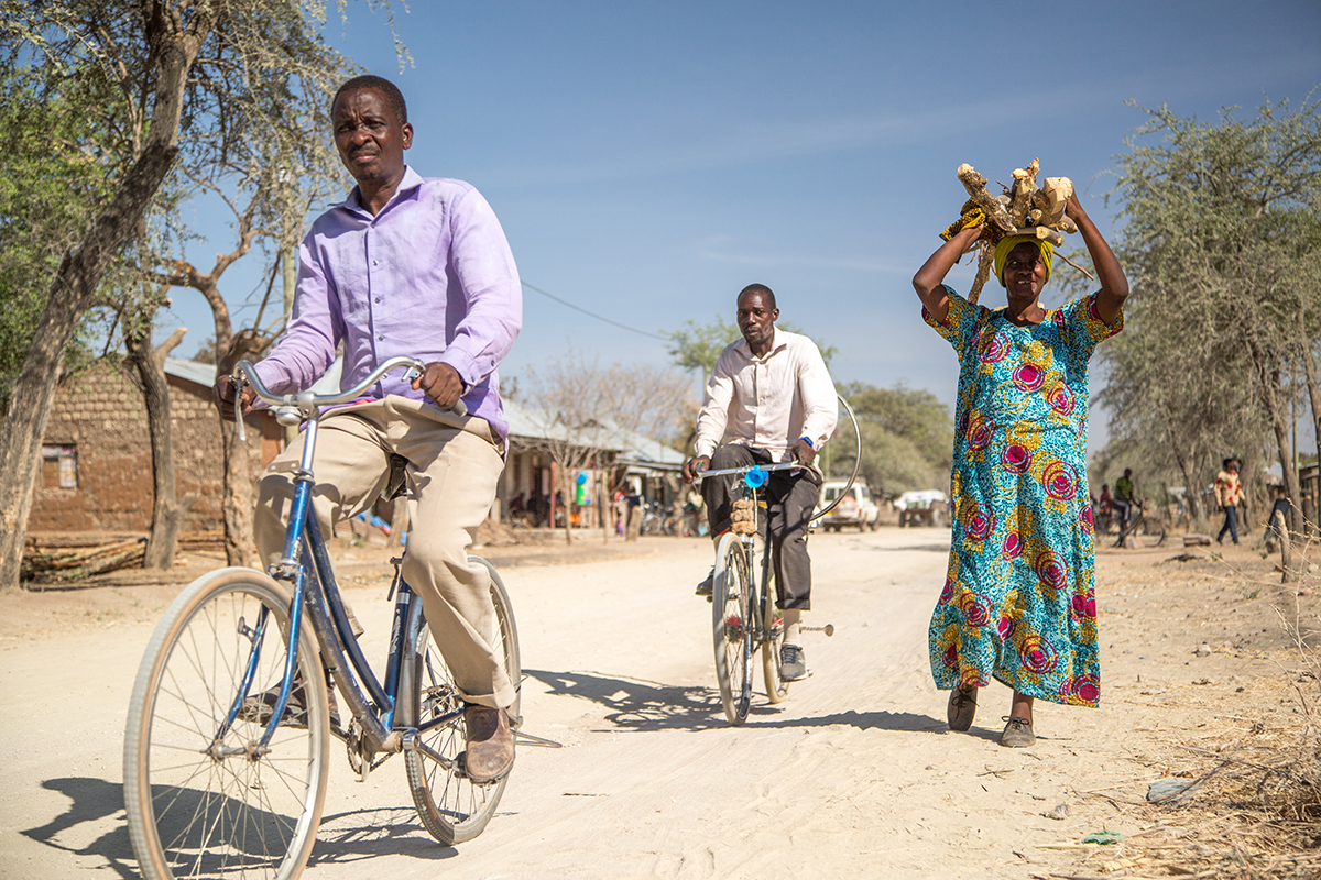 Socha and another man ride bicycles through the village, while Stella walks carrying items above her head.