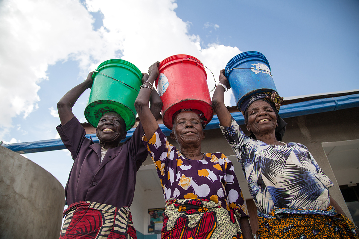 Three women stand smiling with buckets of clean water on their heads.