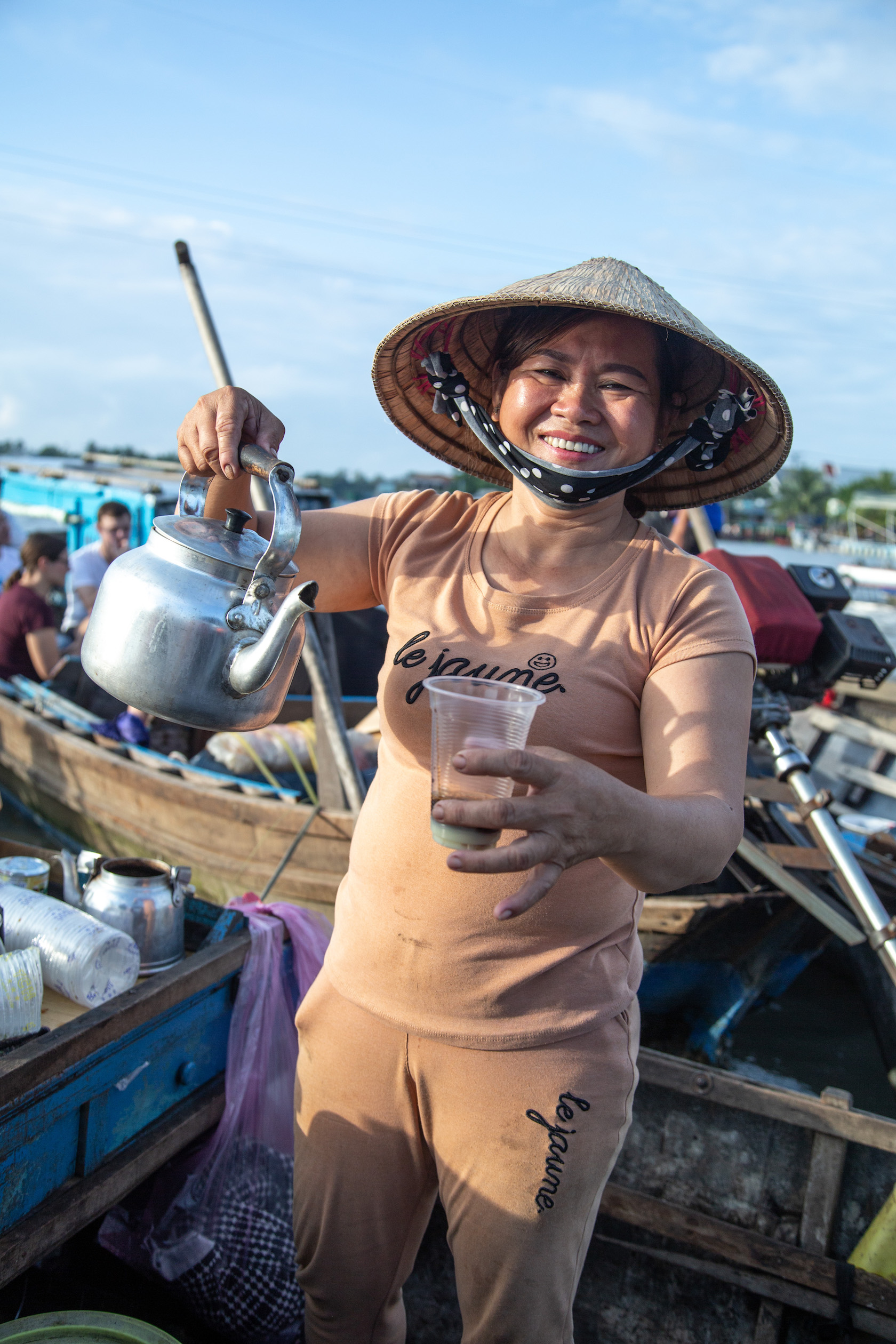 A woman smiles while holding a kettle and a cup of coffee near her boat storefront.