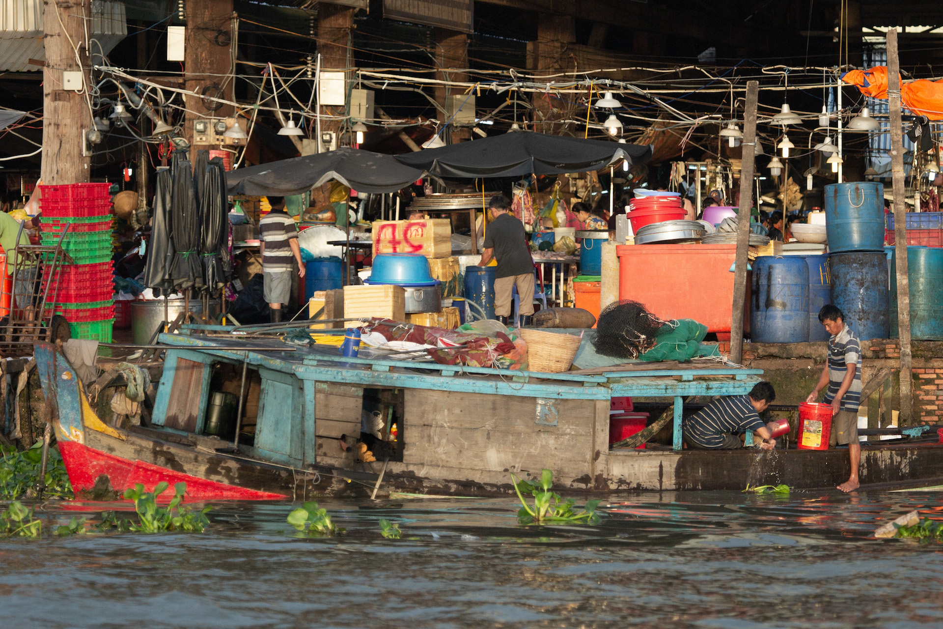 A group of vendors handle their goods with a boat sitting nearby on the water.
