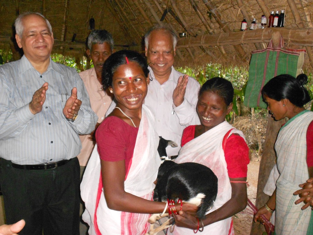 One Heifer India project participant gives a young goat to another recipient as visiting dignitaries look on