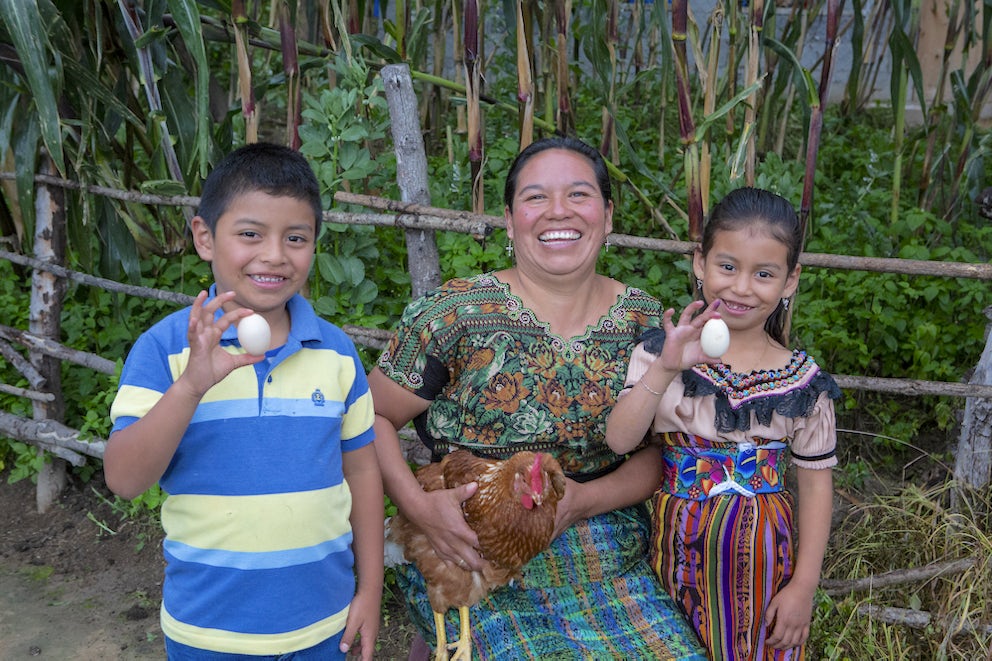 A woman in Guatemala poses with a chicken, alongside two children holding eggs