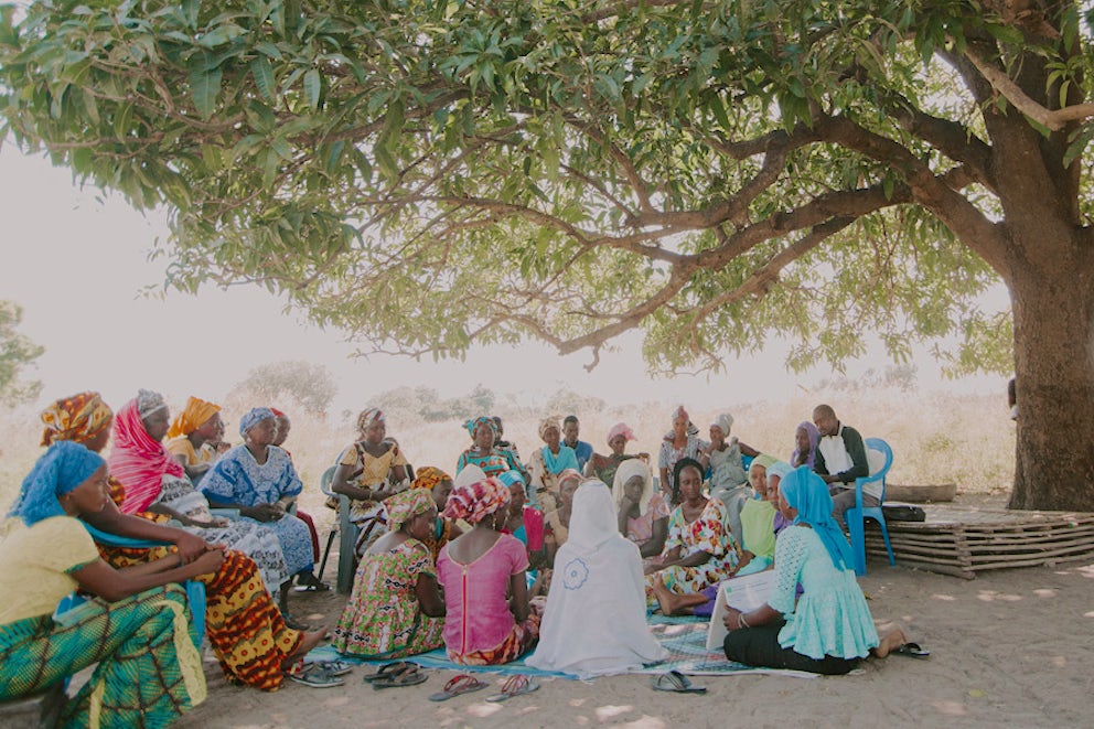 A large gathering of women in colorful, traditional dress sit together on the ground, engaging in what appears to be a meeting.