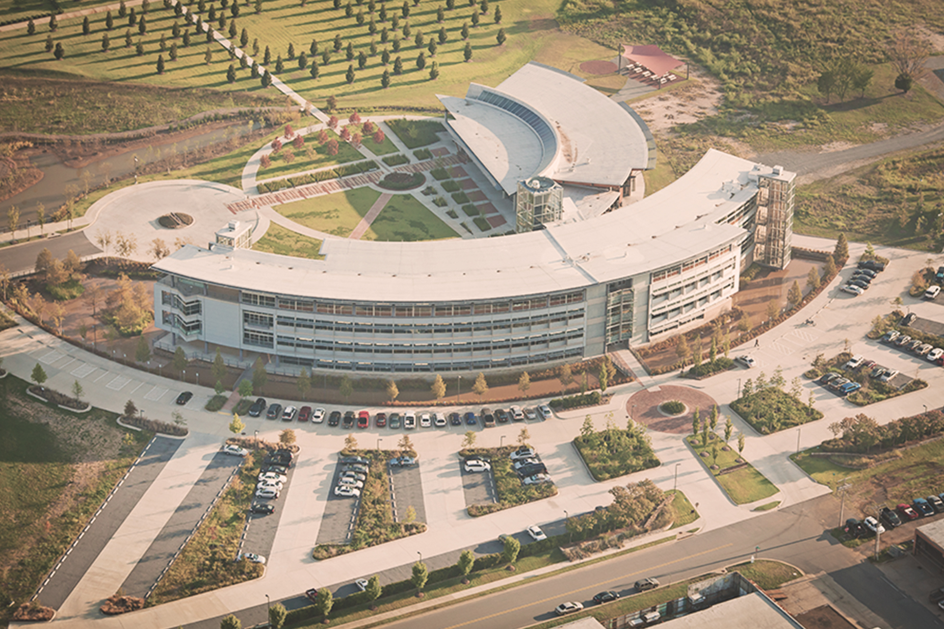 The aerial view of Heifer Headquarters shows how the building is constructed as two crescents, a large one with the main entrance on its bowed side and a smaller one jutting just off the inside of the larger one. The building is surrounded by wetlands with a parking lot out front. 