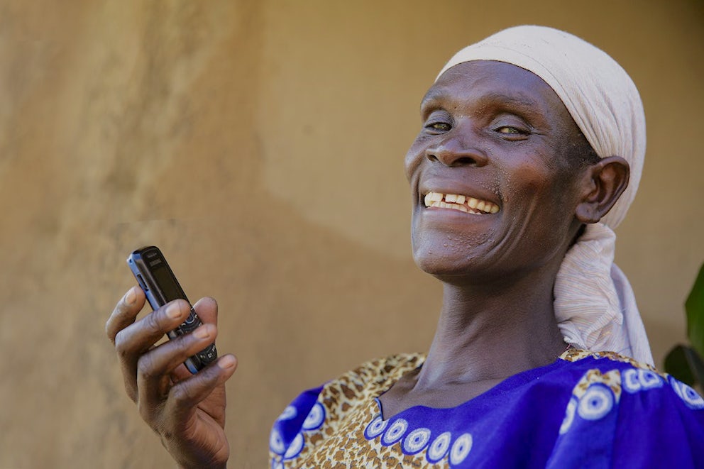 Close-up of woman in desert-like setting smiling at camera and holding a cell phone.
