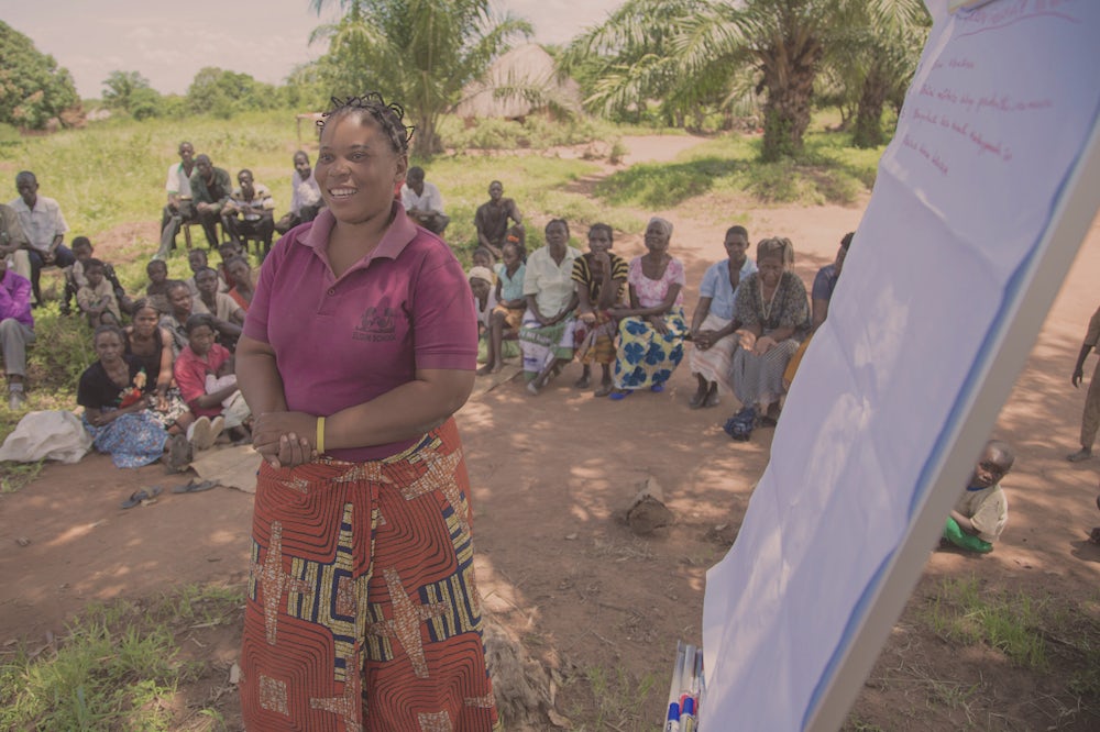 A woman outside in Zambia presents to a group of women sitting around her. Next to her is a large piece of paper propped up with notes scrawled on it.