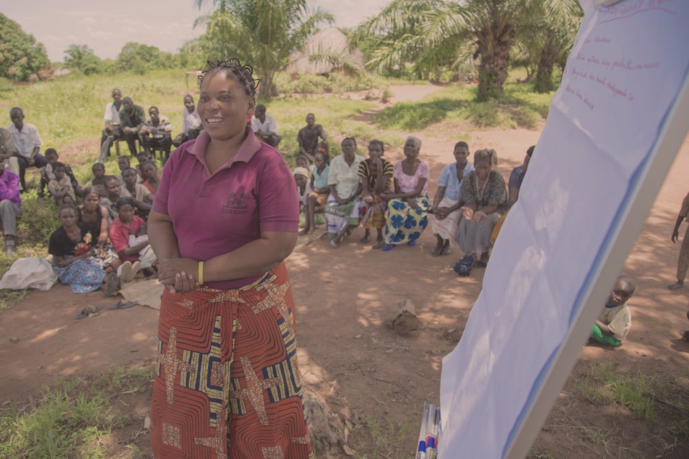 A woman outside in Zambia presents to a group of women sitting around her. Next to her is a large piece of paper propped up with notes scrawled on it. 