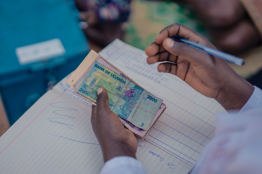 A person with a ledger in their lap holds Ugandan cash in one hand and a pen in the other.