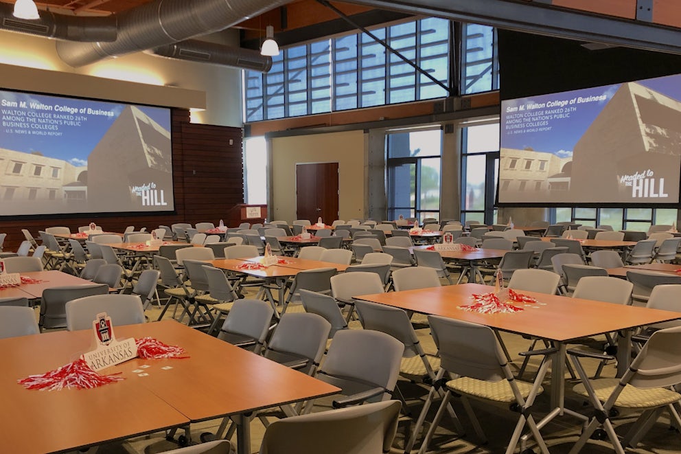 A room is filled with tables topped with pompoms and small signs for the University of Arkansas. Along with natural light flooding in, there are two big screens showing a building that says “Sam M. Walton College of Business: Headed to the Hill.” And a reference to U.S. News & World Report ranking Walton College 26th among the nation’s public colleges.
