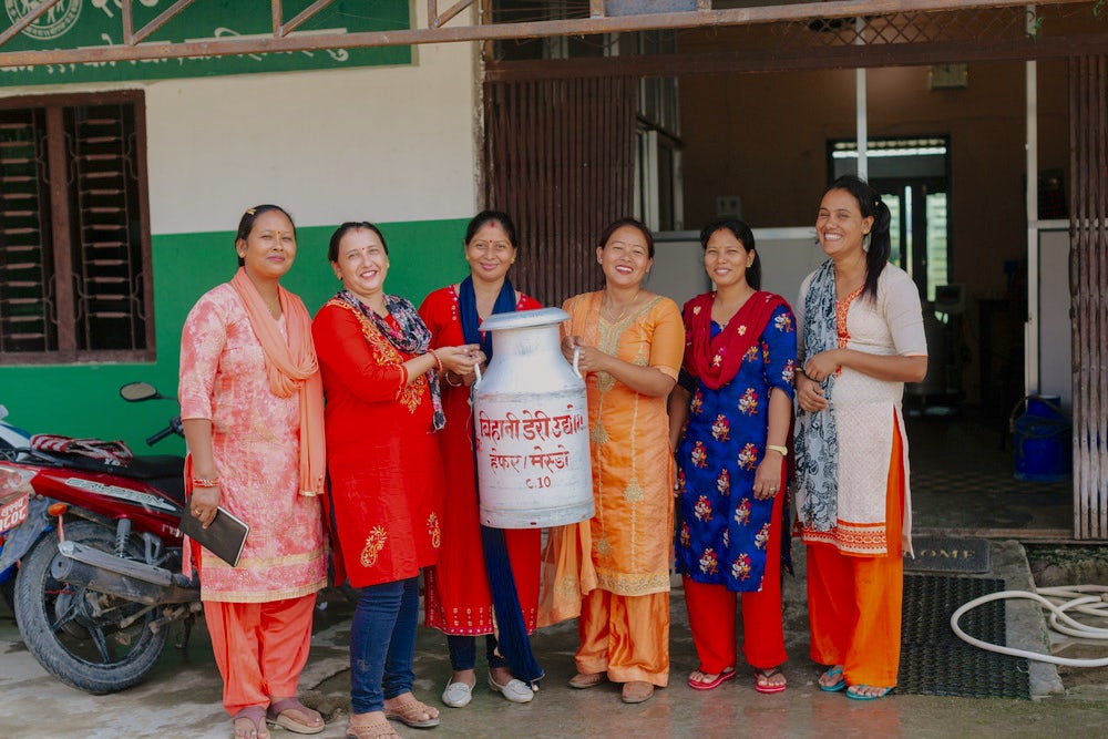Six Nepali women stand next to each other, smiling, and holding a large can of milk.