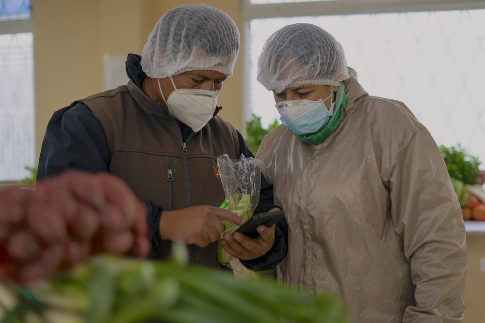 Two people wearing masks and hair nets assemble fruits and vegetables into boxes.