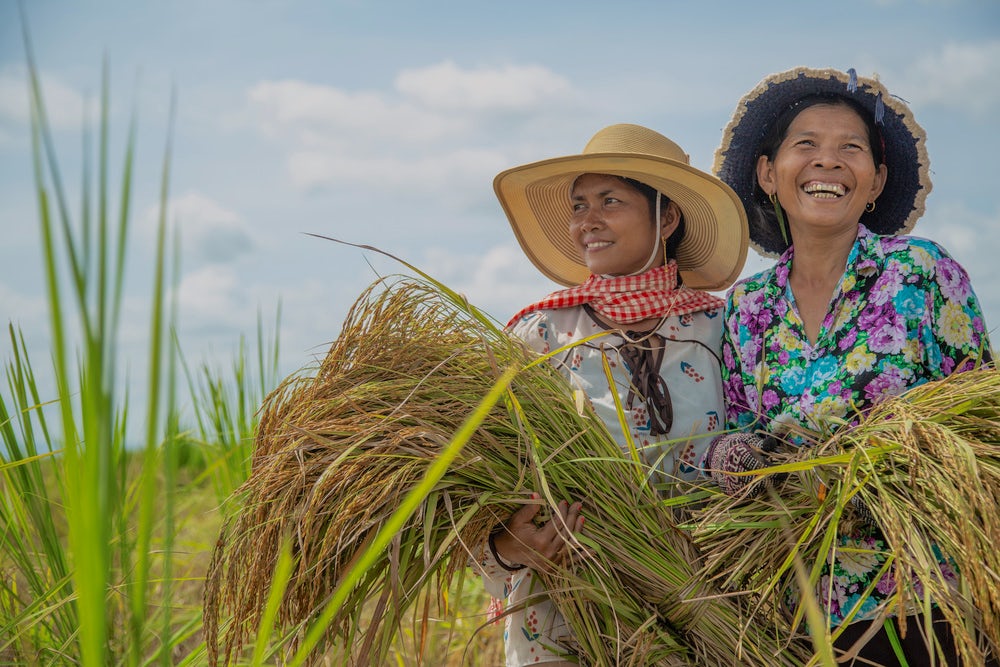Two smiling Cambodian woman stand next to each other, each holding recently-harvested bundles of grass.