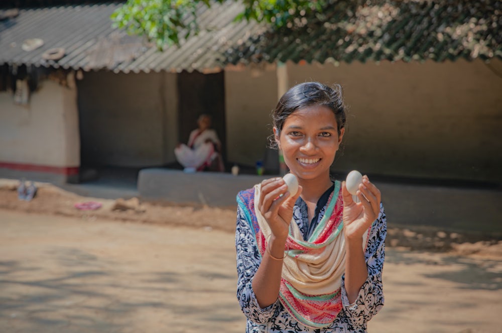 A young Indian woman holds a chicken egg in each hand and smiles at the camera.