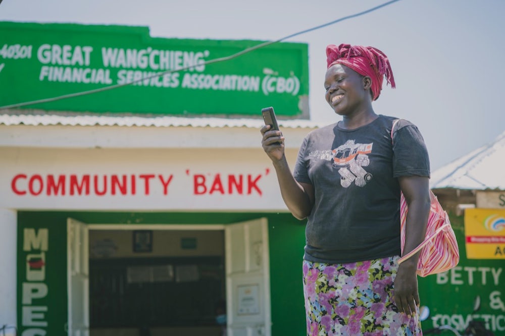 A Kenya woman, wearing a floral skirt, stands in front of a community bank and smiles at the cell phone she holds in her hand.