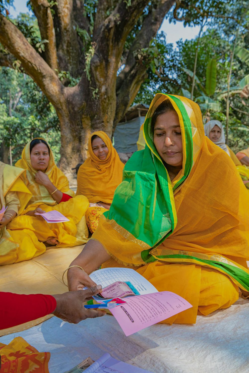 A woman in a yellow and green saree sits on the ground, reading a pamphlet, with other women in yellow sarees in the background.