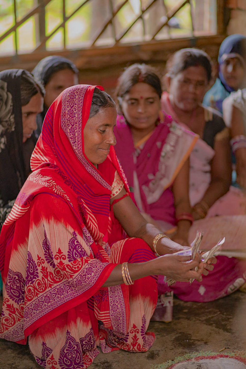 A woman in a red and pink saree counts money, focusing intently, with other women seated around her in a semi-circle.