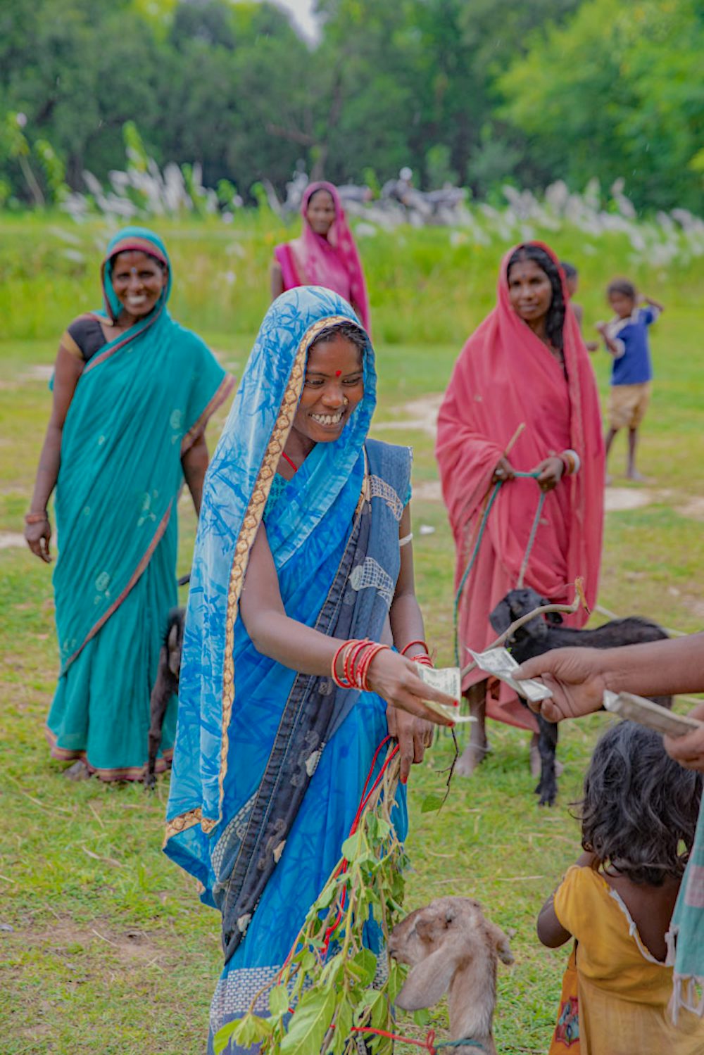 A woman in a blue saree exchanges money in a rural setting with goats and other villagers around her.