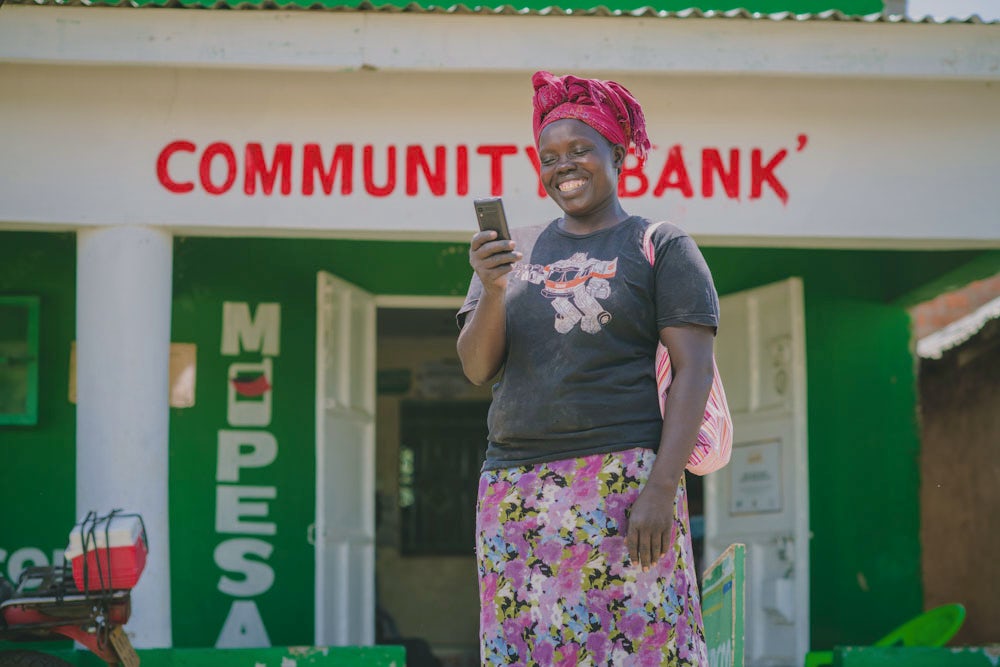 A woman in a printed skirt and red headwrap stands outside a community bank holding a mobile phone.