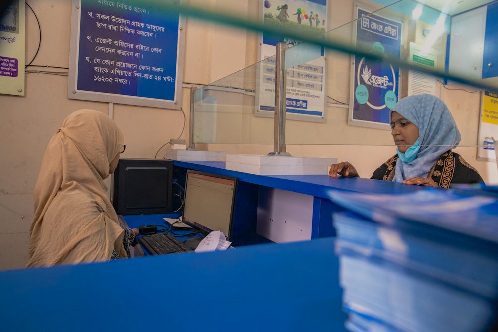A woman stands at the desk of a bank teller in Bangladesh.