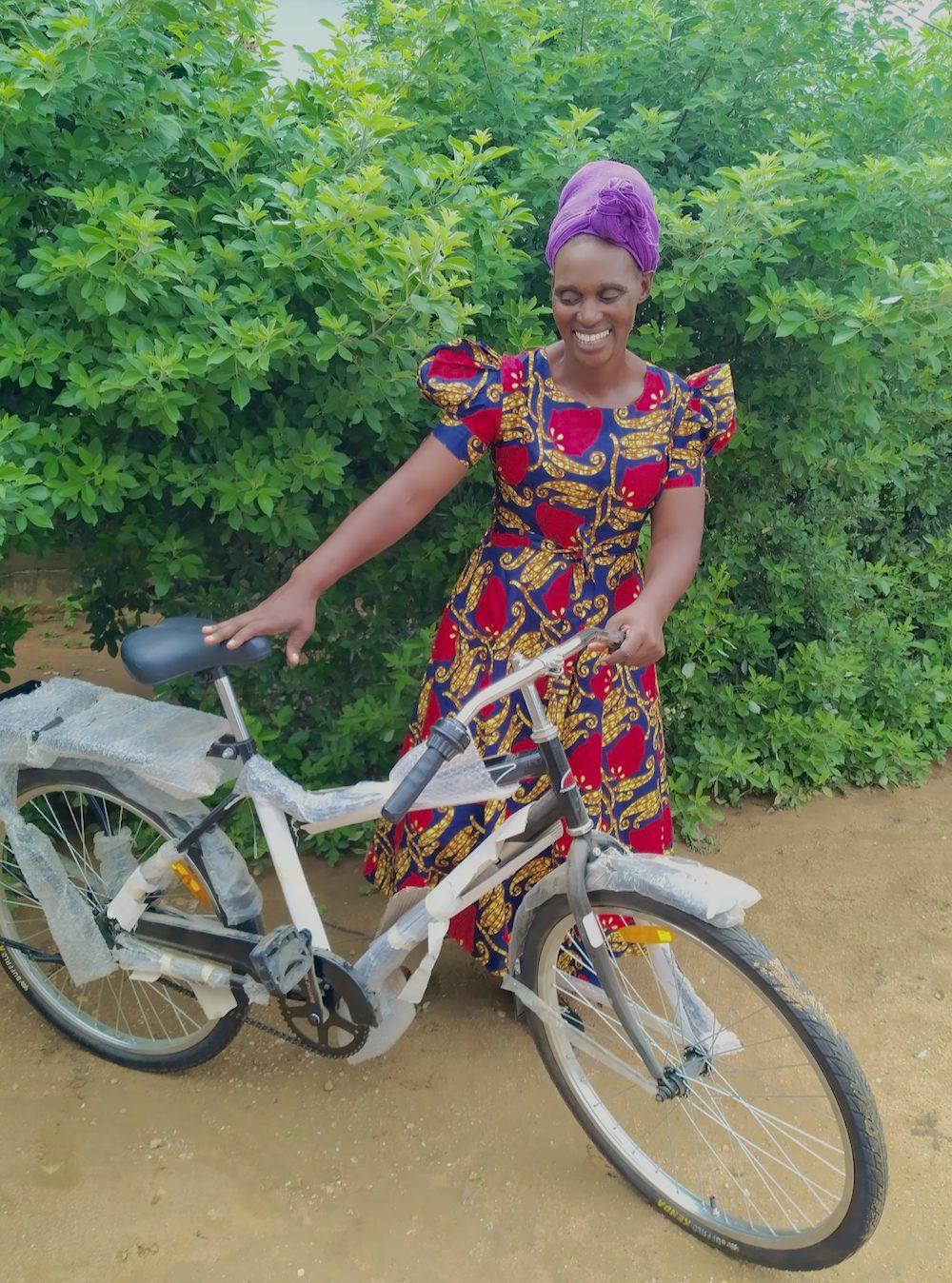 A smiling woman wearing a purple headscarf and floral dress admires her new bicycle.