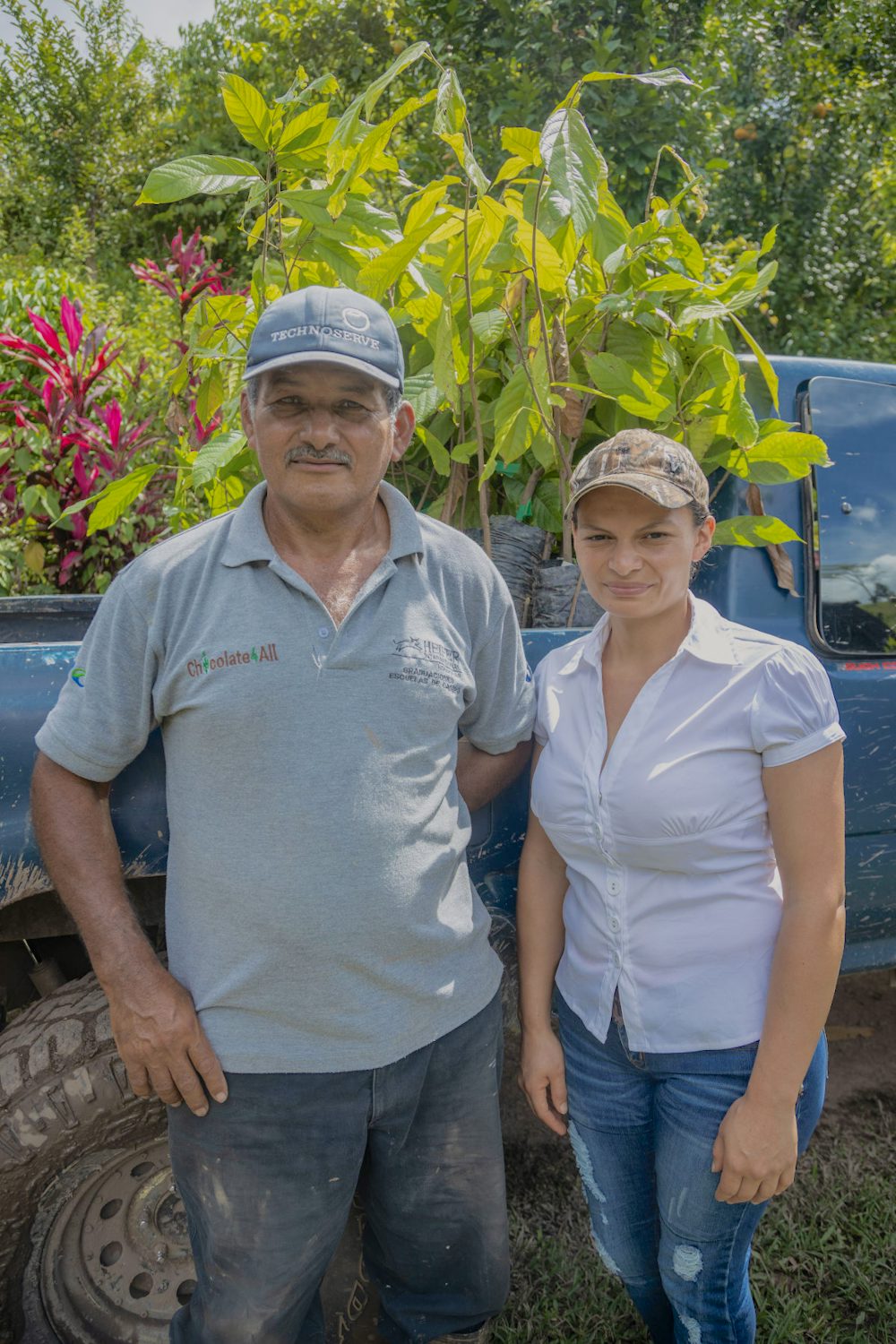A young woman and a man standing in front of a truck of cacao plants they grow in their farm. 