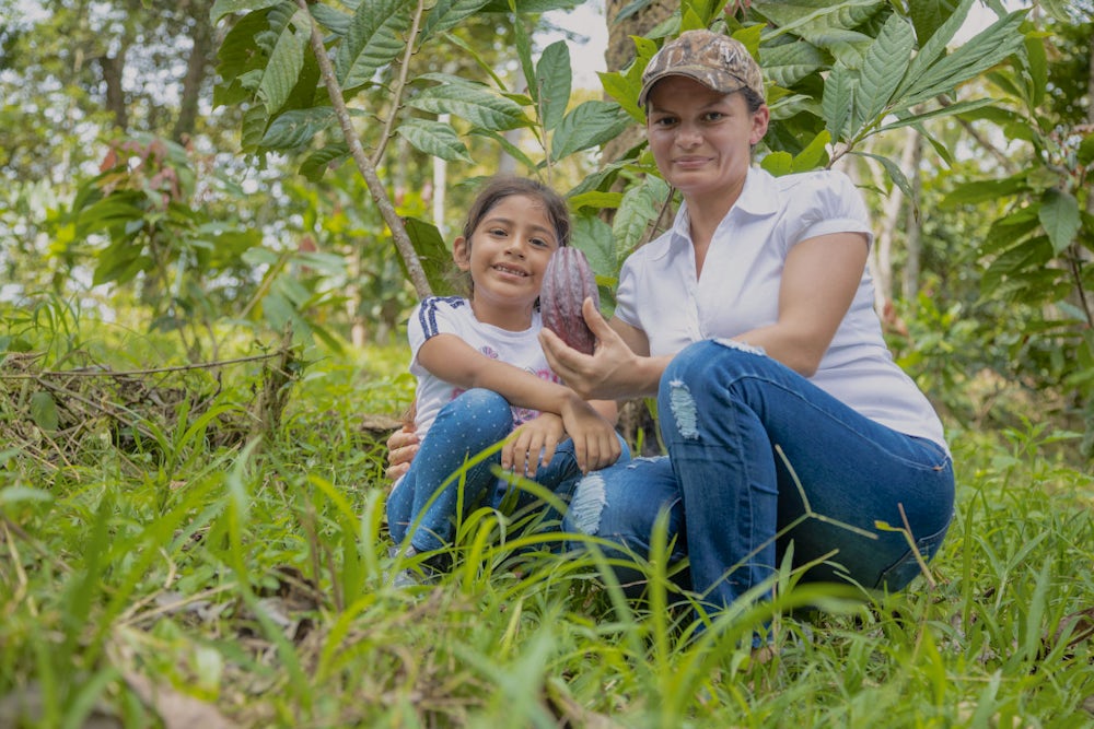 A young woman sitting beside her daughter, while holding a cacao plant in her hand. 