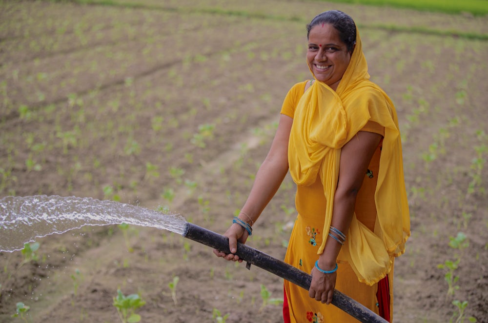 A woman wearing a yellow dress and scarf waters her field with a hose.