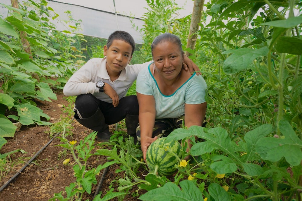 A woman and her son crouch next to a large watermelon in their greenhouse.