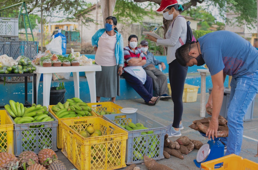 A shot of a local market shows people weighing produce and looking at fruits and vegetables displayed on tables.