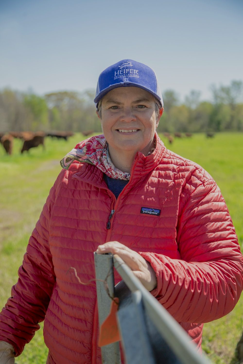 A woman standing in a field holds a gate with her left hand and smiles at the camera.