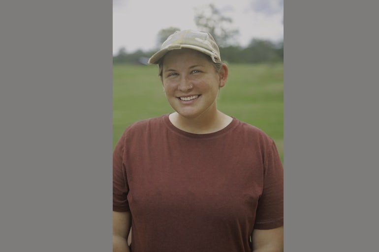 A woman wearing a red shirt and a baseball cap smiles at the camera.