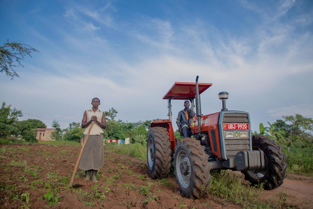 A woman holding a hoe and standing beside a tractor being driven by her husband in their field.