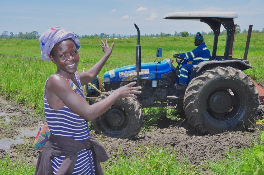 A woman stands in front of a tractor, overseeing another person operating it in an agricultural setting.