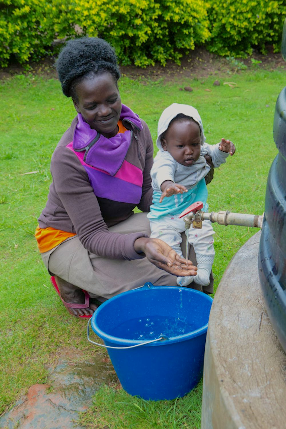 A woman holding her child gathers water from a rainwater collection tank into a blue bucket.