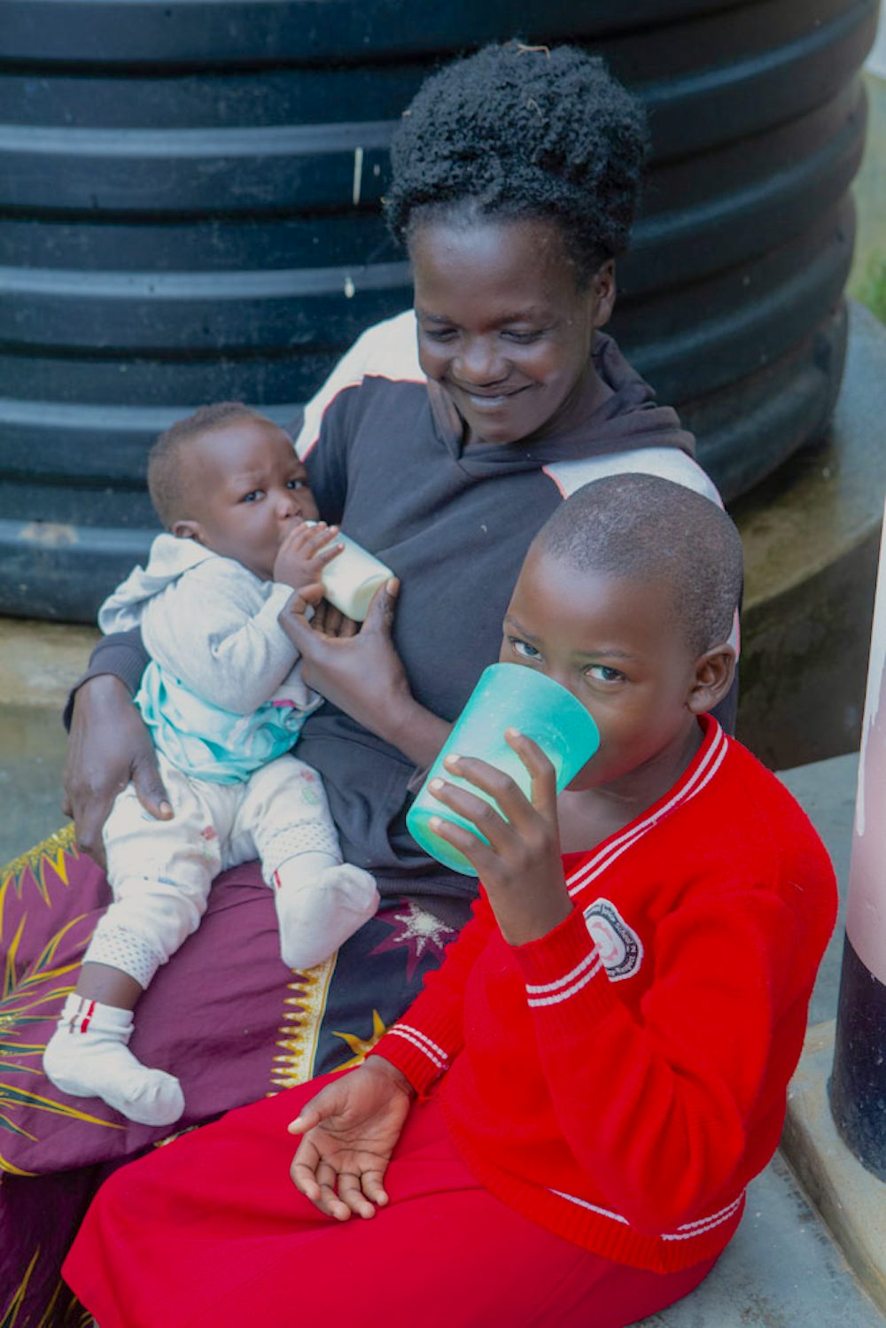 A woman on a step cradles a toddler while another child drinks from a cup.