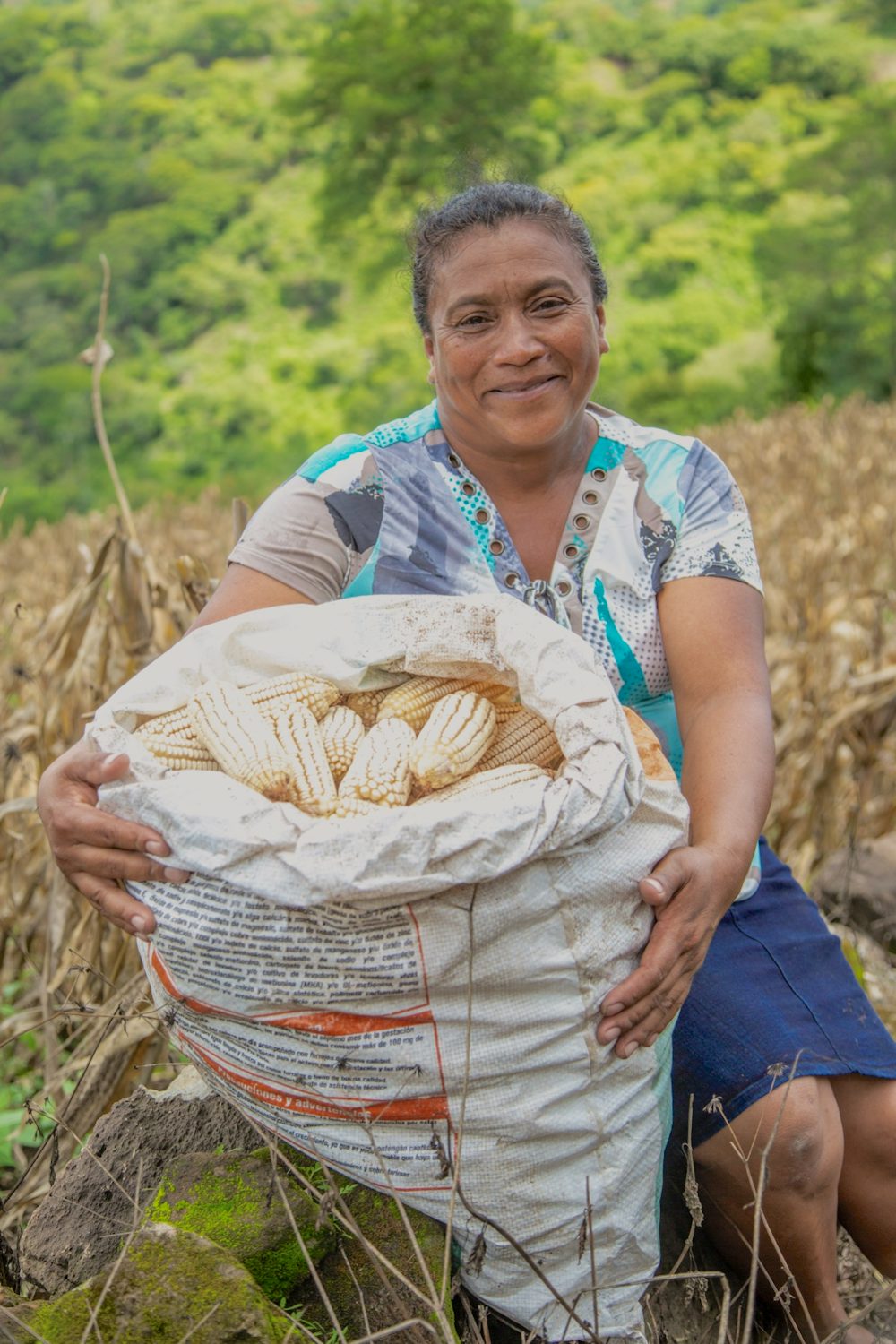 A woman sits in a field, holding a large bag of harvested corn.