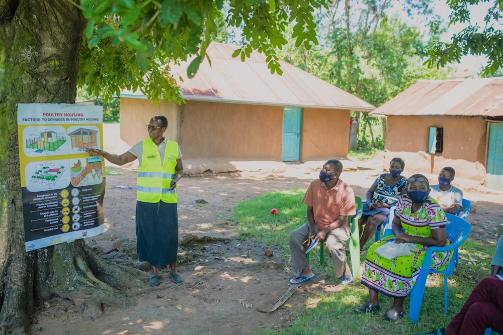 A woman conducting a training with the help of a chart in an open ground with several farmers seated on chairs.
