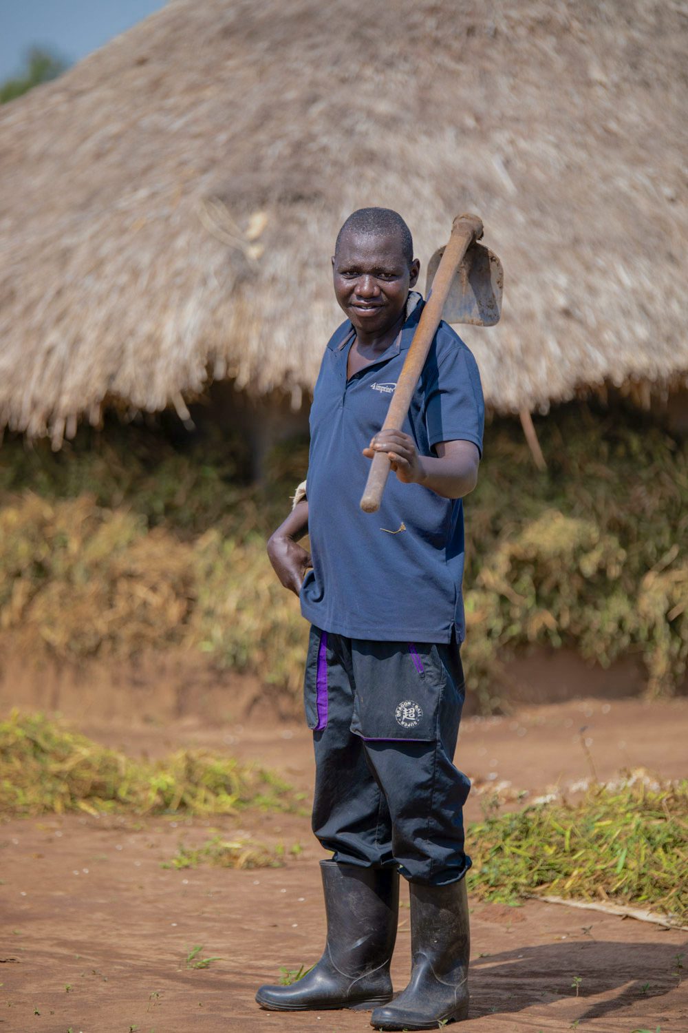 A farmer stands proudly before a thatched-roof hut, holding a hoe over his shoulder.