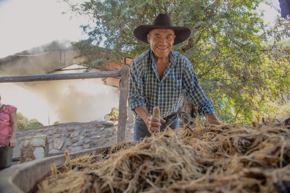 A man uses a stick to stir a pot of pulp as he smiles at the camera.