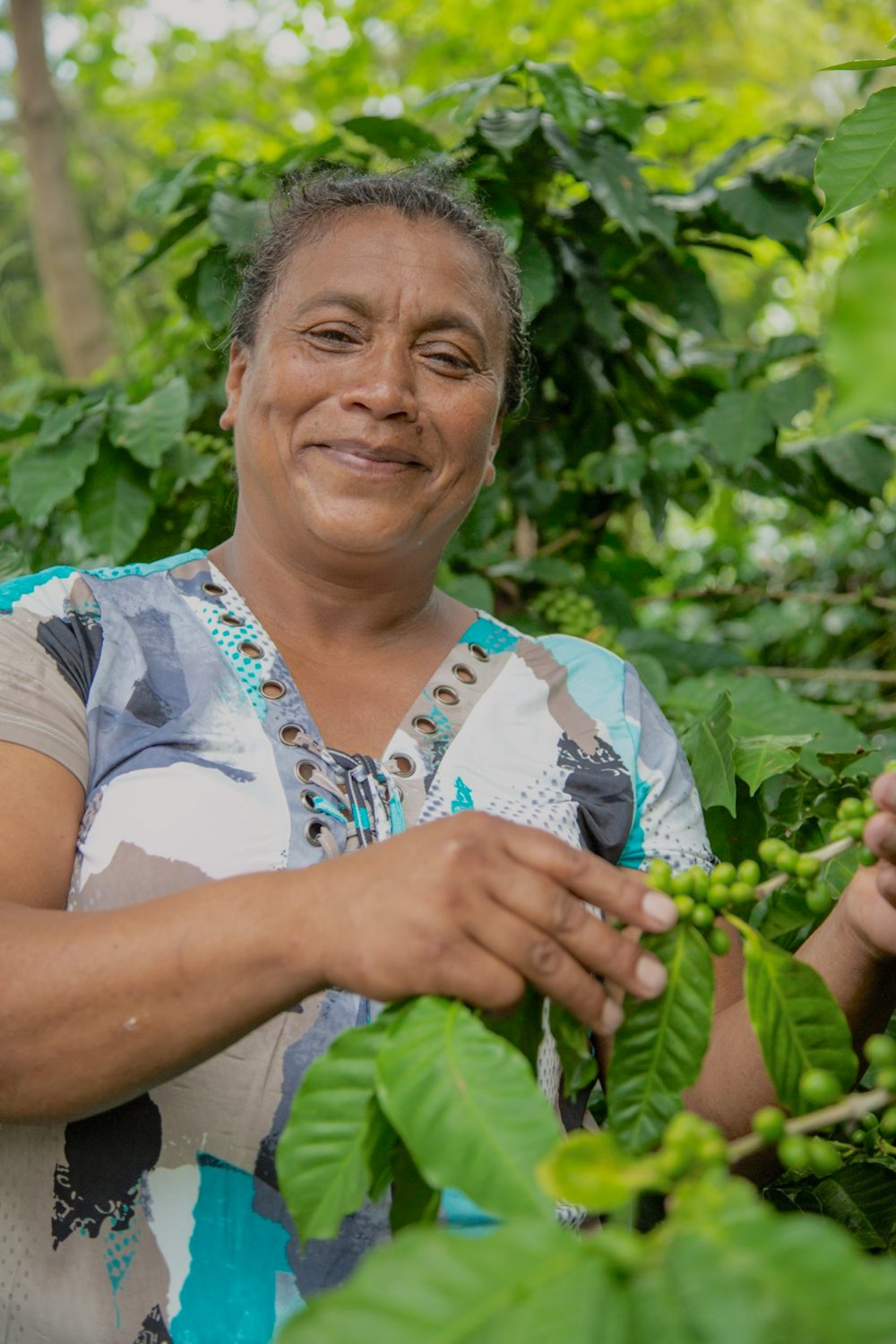 A woman stands next to her coffee plant and smiles at the camera.